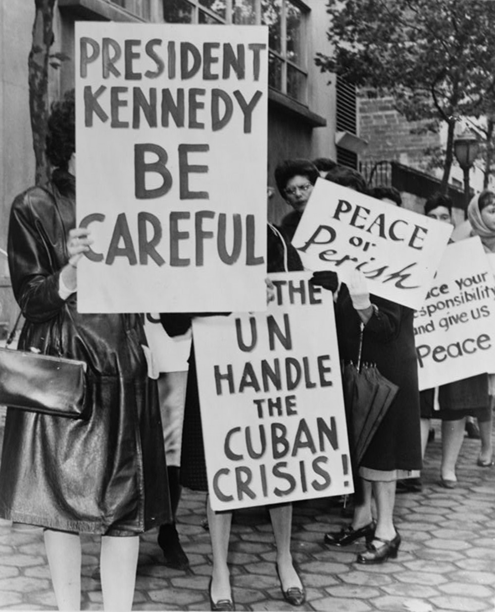 Protestors hold signs that read "President Kennedy Be Careful," "Let the UN Handle the Cuban Crisis!," "Peace or Perish," and "[unclear] your responsibility and give us peace."