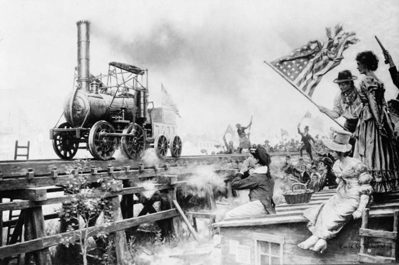 People wave an American flag as they watch a train engine move along tracks.