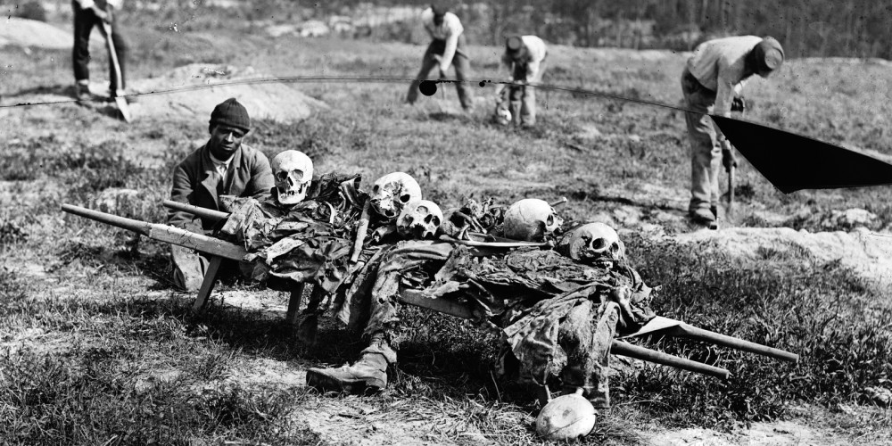 A young man by a cart that holds several human skulls and other body parts.