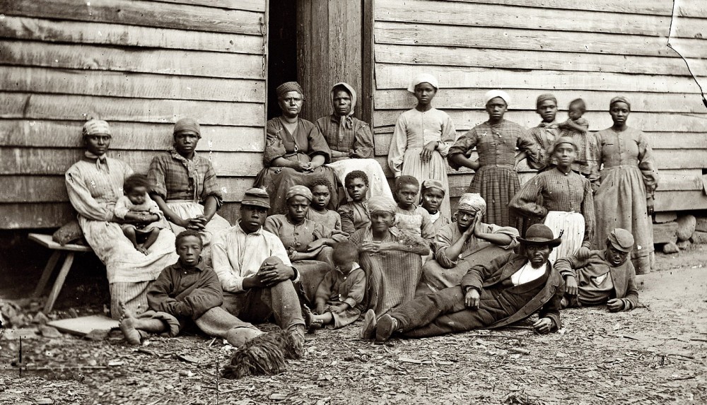 This photograph shows twenty three men, women, and children sitting together in a contraband camp. 