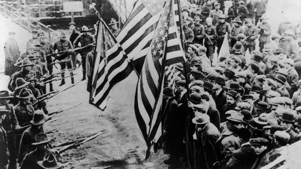 This photograph shows striking textile workers carrying American flags facing off against soldiers with bayonets affixed to their rifles. 