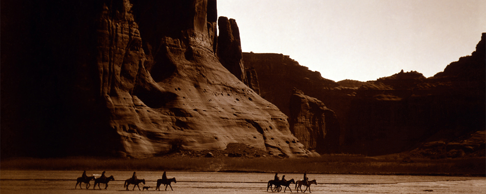 Photograph of Navajo riders on horseback through a desolate canyon. 