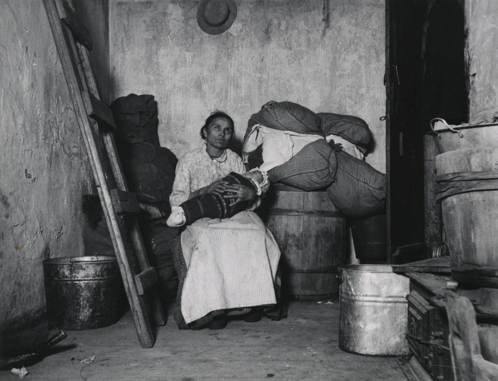 Photograph of a woman sitting in a dirty room and holding a baby. 