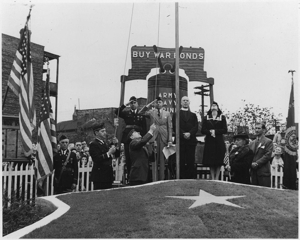 Photograph of a fundraising event selling war bonds. Soldiers salute as a flag is raised. 