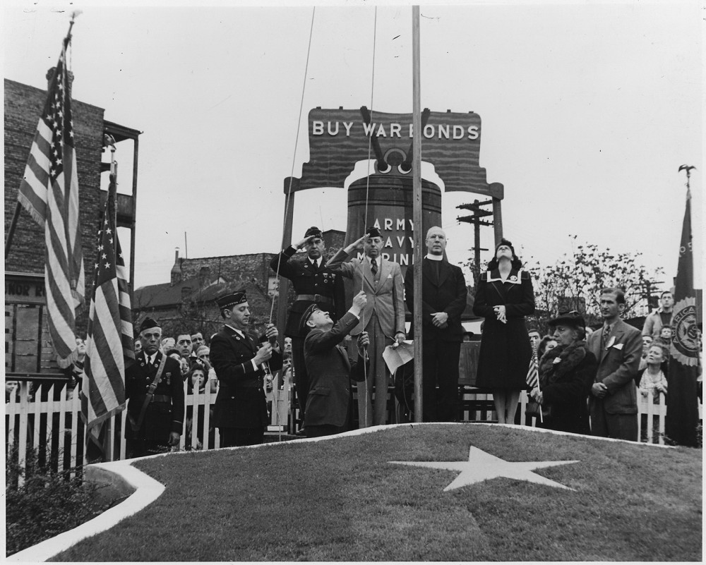 Photograph of a fundraising event selling war bonds. Soldiers salute as a flag is raised.