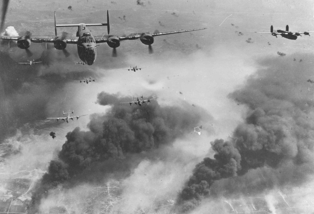 Photograph of several Allied bombers in flight. Dark smoke rises from the ground below. 