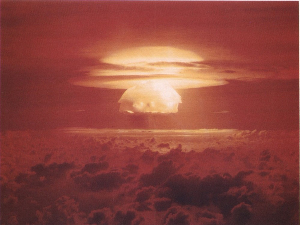 Photograph of a nuclear weapons test. A mushroom cloud rises above the clouds. 