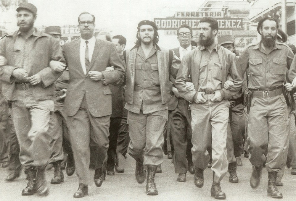 Photograph of revolutionaries, including Che Guevara and Fidel Castro, marching arm and arm. 