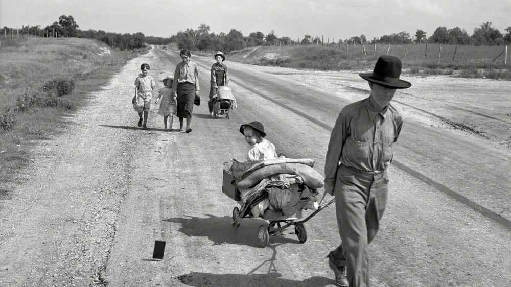 Photograph of a family traveling by foot down a road. A small child is in a wheel barrow packed with blankets. 