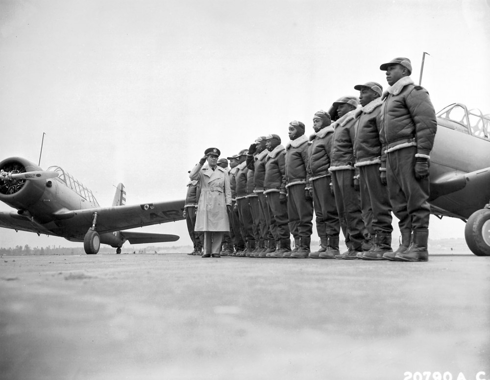 Photograph of several Tuskegee Airmen standing at attention. Their commanding officer salutes. A fighter plane is in the background.