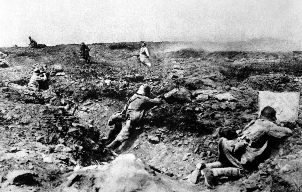Photograph of soldiers crouching behind objects in a desolate, charred landscape. 
