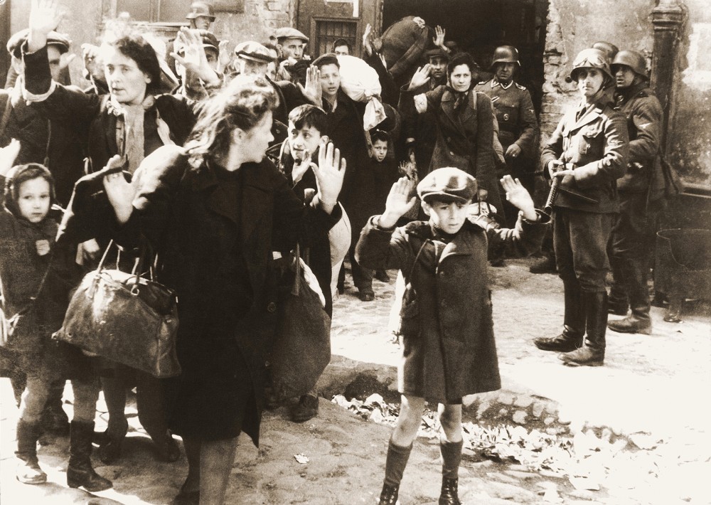 This photograph shows a number of Jewish women and children, including a small boy in the foreground, with their hands raised in surrender while German soldiers keep their weapons trained on the civilians.