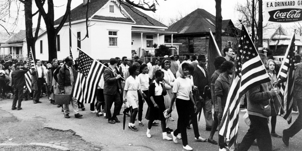 Photograph of civil rights protestors marching from Selma to Montgomery. Many are carrying American flags. 