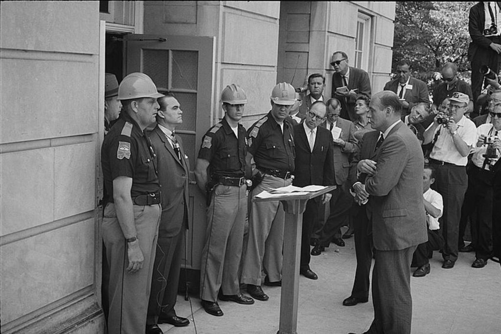 A man stands in front of a door, flanked by law enforcement officers, as another man stands before him.