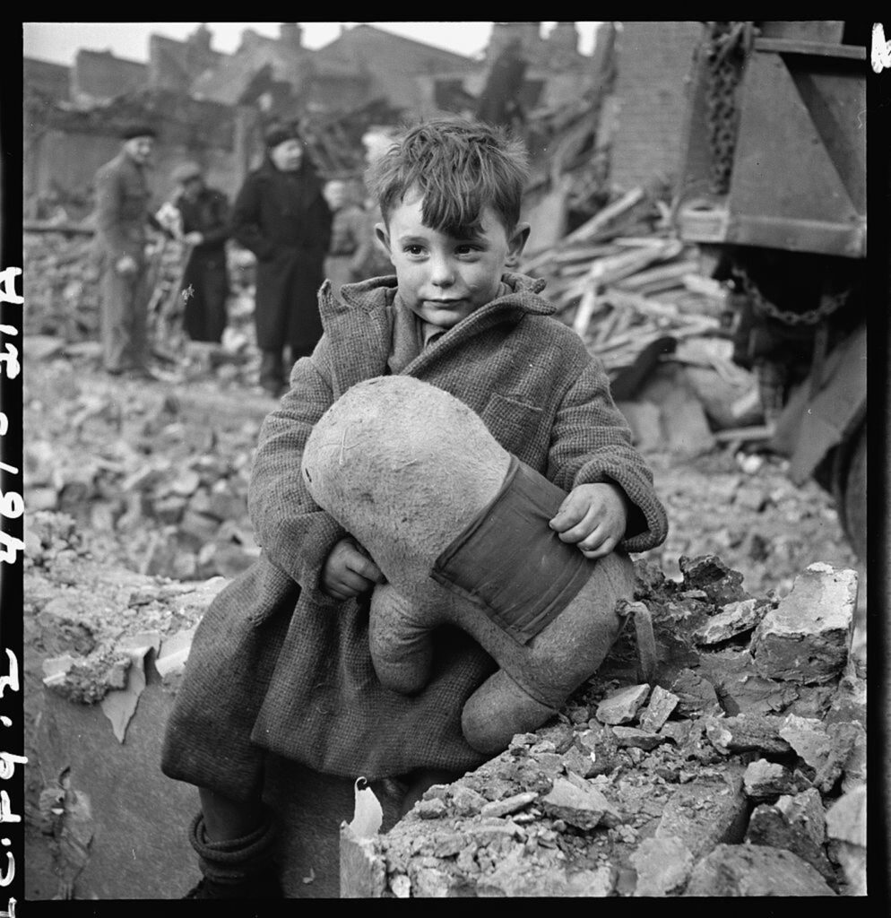A child, holding a stuffed toy, sits among rubble.