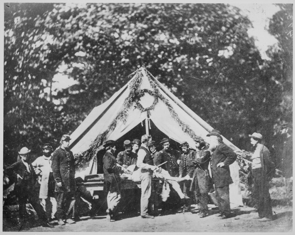 Photograph of a medical tent with about a dozen men huddled around a body on a table. A man in the foreground holds what appears to be a saw or knife. “Amputation being performed in a hospital tent, Gettysburg,” July 1863. National Archives and Records Administration.
