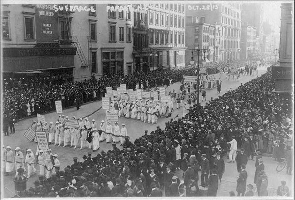 Women dressed in white march in a parade with throngs of people crowded on both sides of the street. 