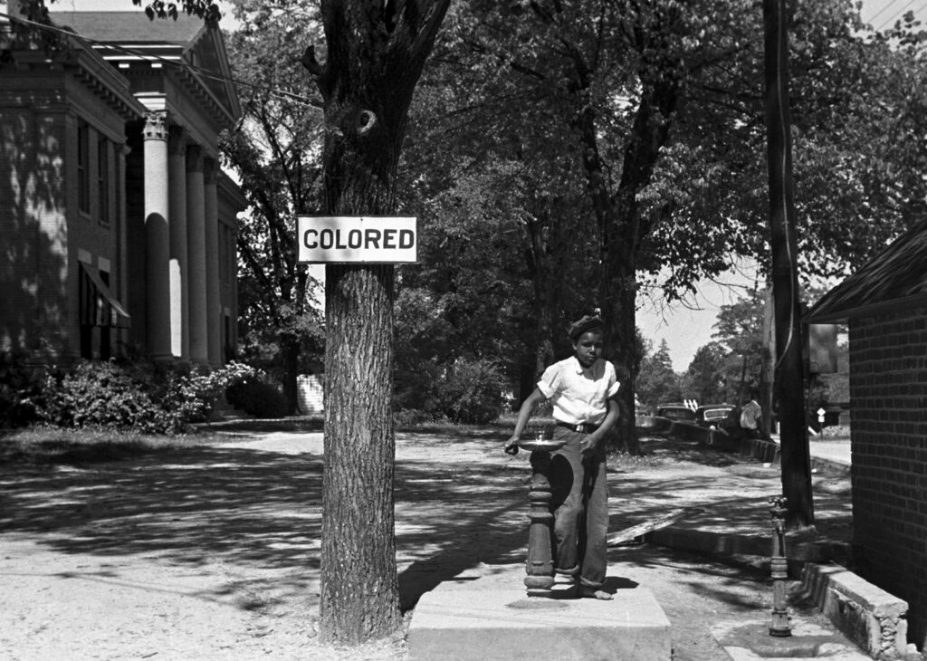 A boy drinks from an outdoor water fountain in front of a tree with a sign reading “Colored.”