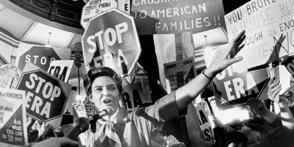 A woman speaks before a microphone as demonstrators hold signs reading “Stop ERA” behind her.