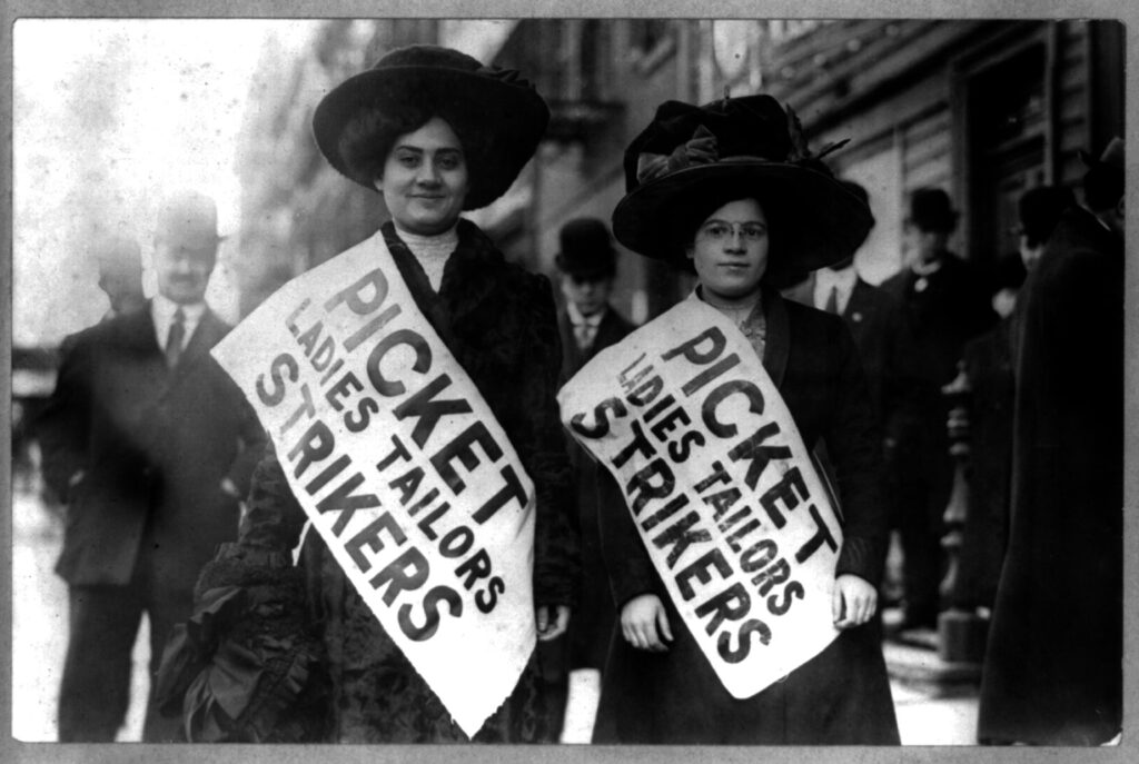 Two women strikers wearing bonnets pose with large sashes reading “Picket Ladies Tailors Strikers.”
