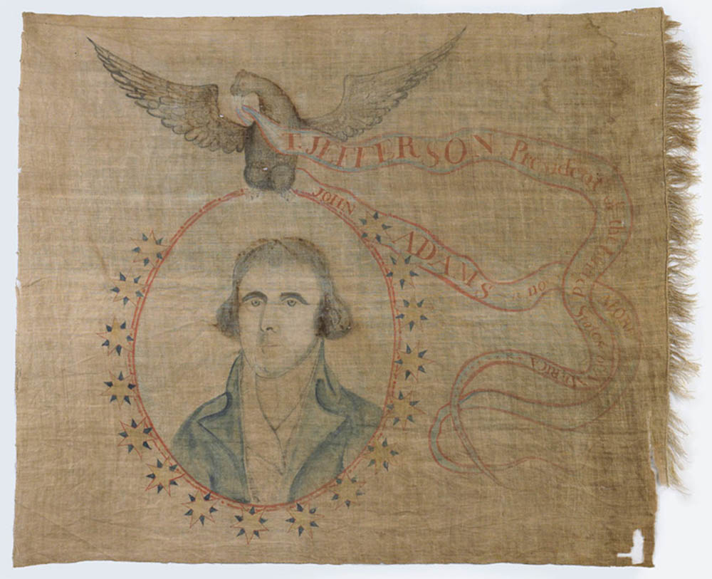 This stitched victory banner celebrating the presidential election of Thomas Jefferson depicts a portrait of Jefferson in the center of a red circle that is outlined by yellow and blue stars. An eagle sits atop the circle with a ribbon in its mouth that reads, “T. Jefferson, President of the United States of America. John Adams is no more.”