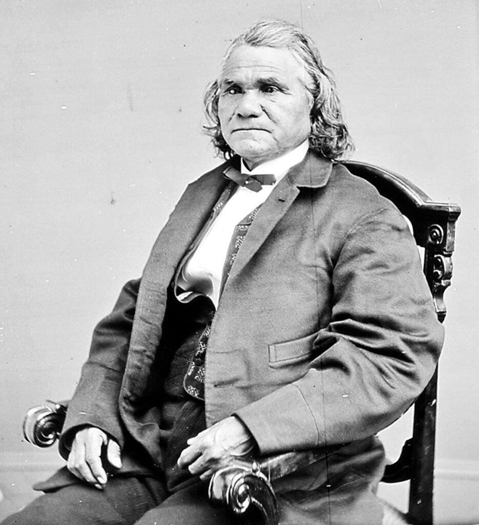 A photograph of Isaac Stand Watie sitting in a chair and looking slightly to his left. He wears pants, dress shirt, vest, and coat with a bowtie. Isaac Stand Watie Degatag, Principal Cherokee Chief, was the last Confederate general to surrender in the Civil War. Wikipedia.