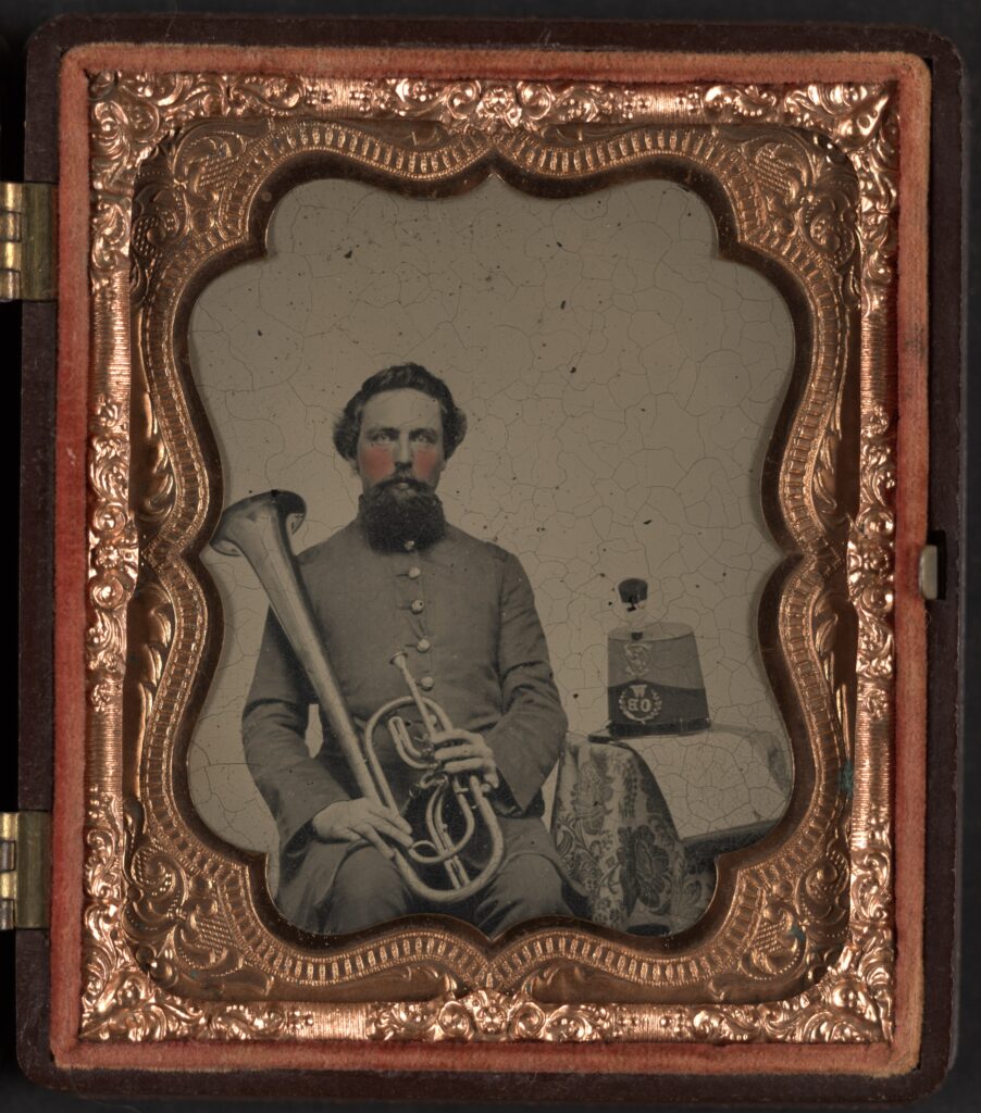 A soldier clad in gray holds an instrument that looks like a french horn. The photograph is placed as a cover to a book that is ornately bound with red trim. Unidentified soldier of Co. H, 13th Virginia Infantry Regiment in uniform with over-the-shoulder saxhorn. Wikipedia.
