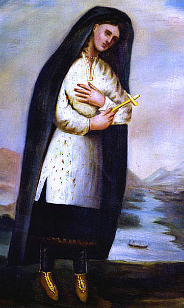 Religious painting of a Native American woman, identified as Kateri Tekakwitha, standing beside a river with mountains in the background. She wears a white tunic adorned with small decorative patterns, a dark cloak over her head and shoulders, dark leggings, and moccasins. Her head is tilted gently to the side in a contemplative pose, and she holds a wooden cross close to her chest with both hands. The setting suggests a serene, spiritual landscape with a canoe or boat on the river below. 