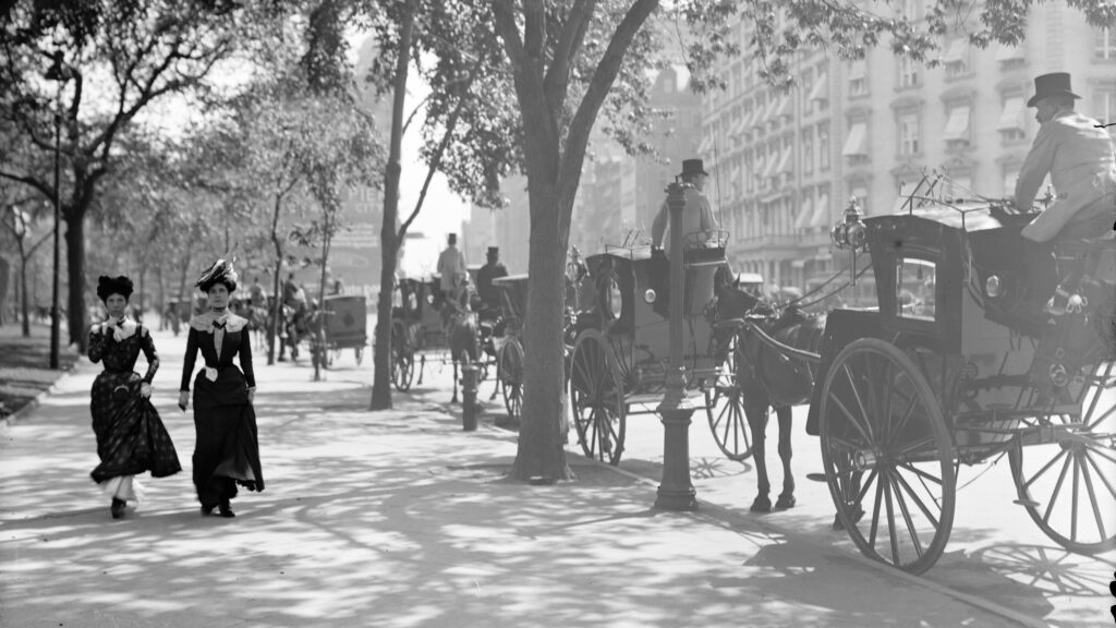 Two women pass by a line of horse-drawn carriages.