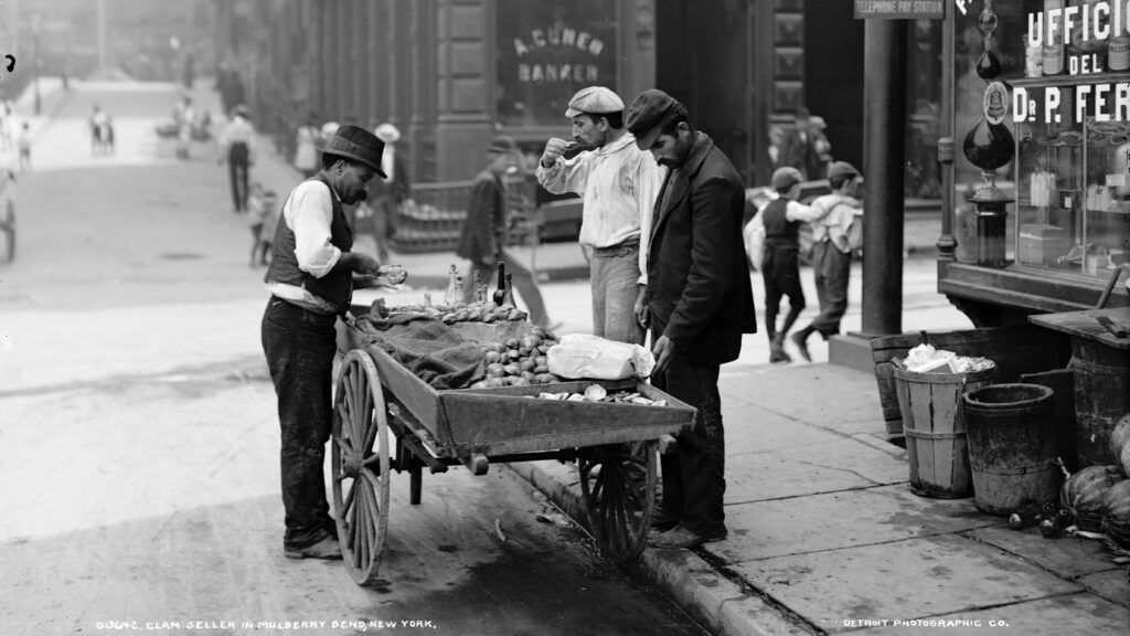 A man sells clams from a cart in an urban setting.