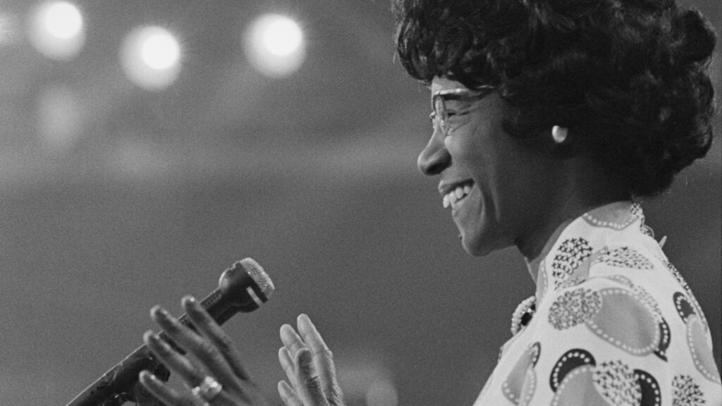 A woman, depicting in a side profile, smiles before a microphone.