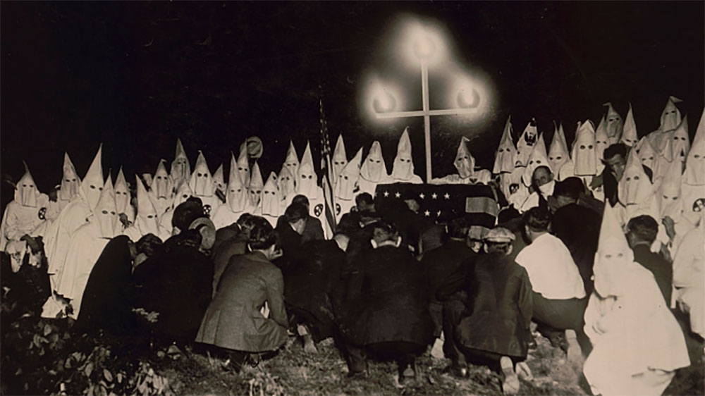 Men in Ku Klux Klan regalia pose for photographers under a cross.