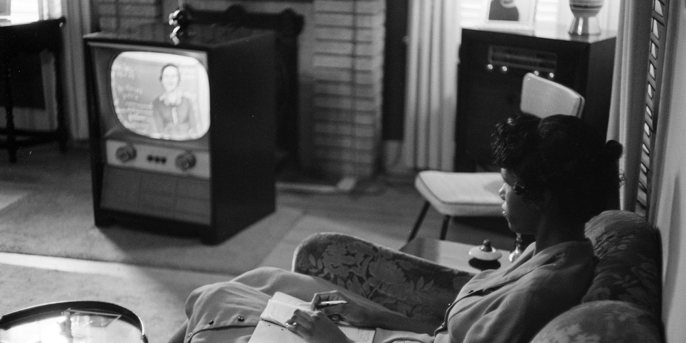 A young woman sits watching a woman in front of a chalkboard on television.