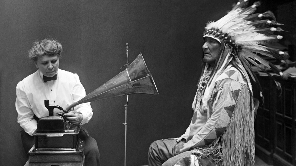 An old man in traditional Indigenous clothing stands before an audio device tended by a woman in a shirtwaist.